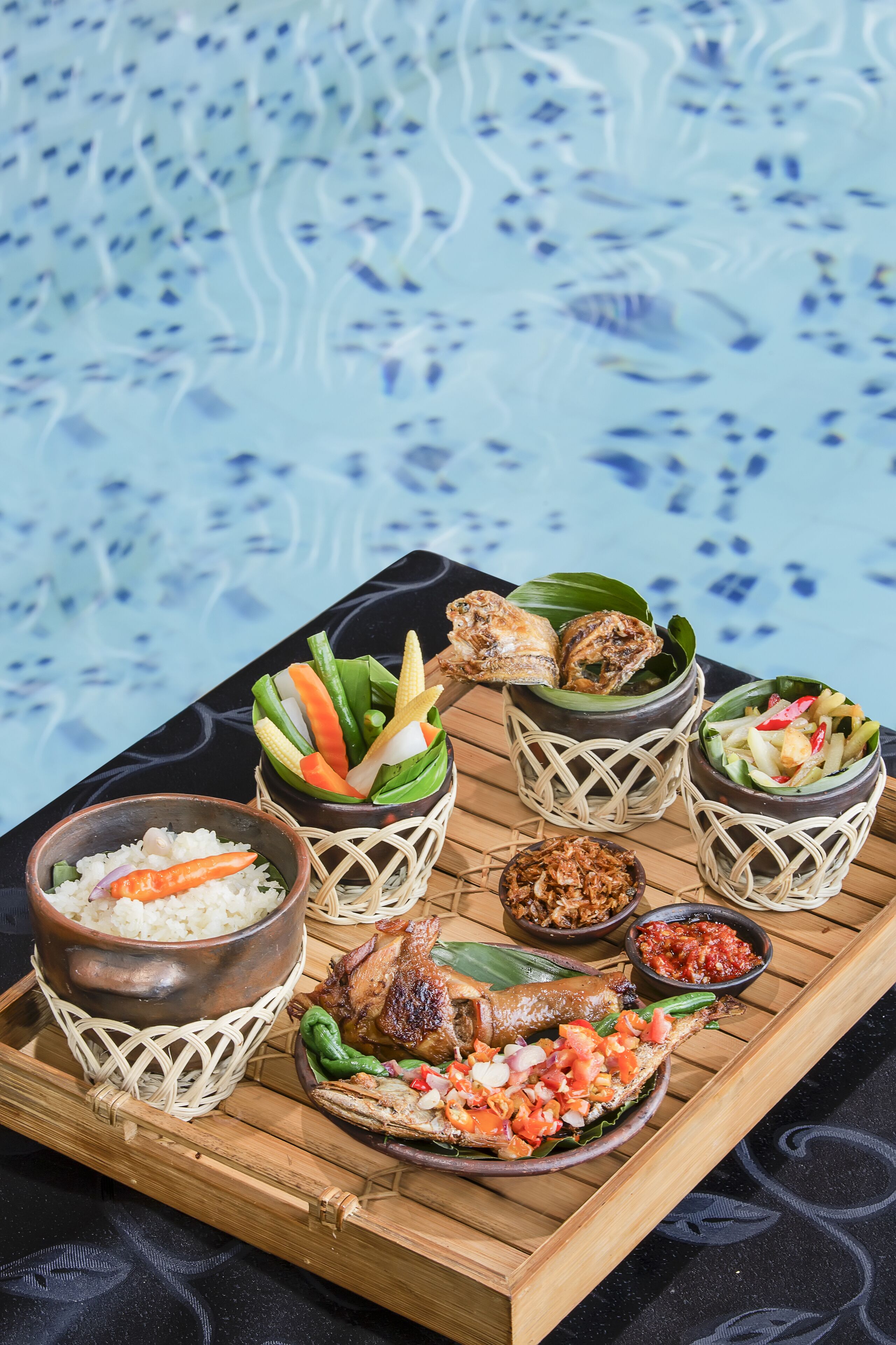 A tray of food with rice, vegetables, and a fish dish, placed on a wooden table by a pool.