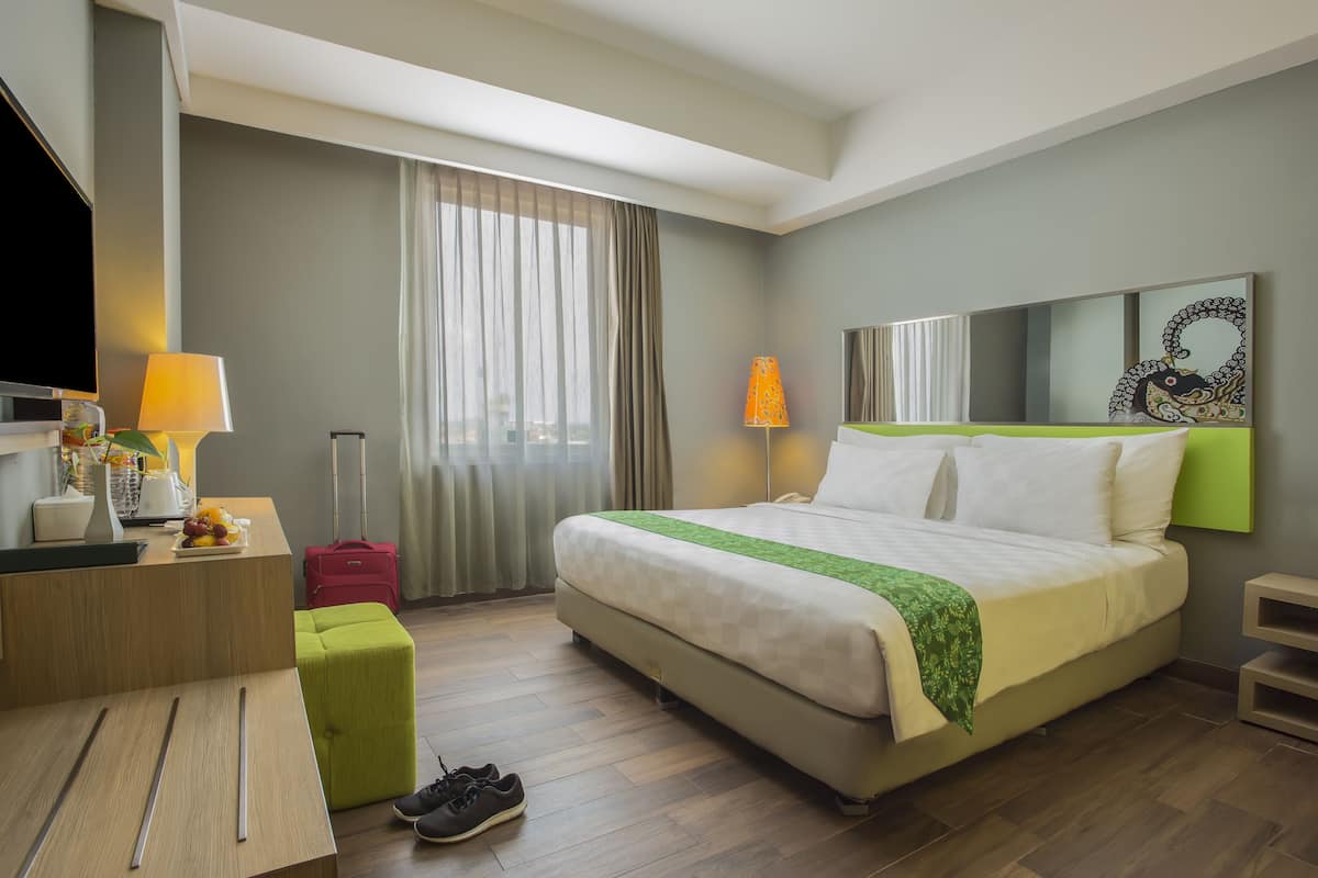 A hotel room with a bed, a desk, a TV, a lamp, a suitcase, and a green ottoman.