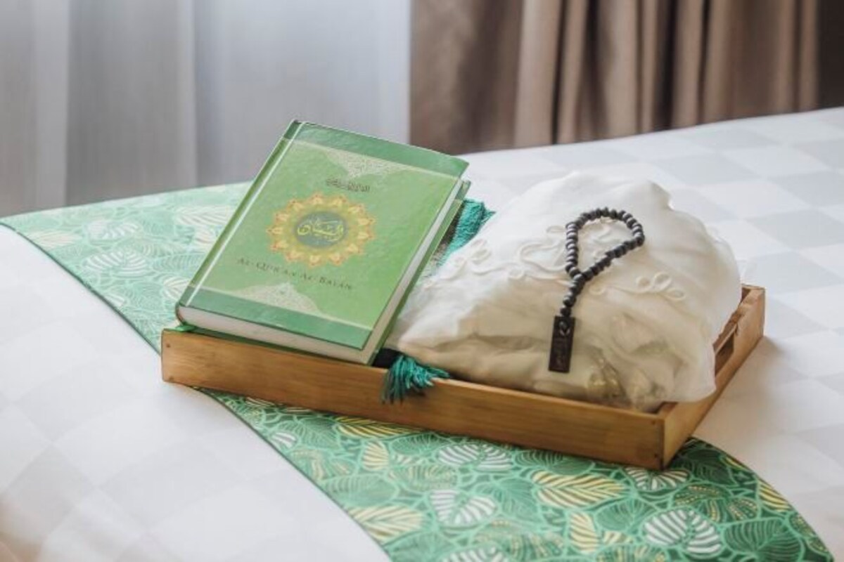 Una cama con una colcha verde estampada, una bandeja de madera con un libro y un paño blanco con un collar de cuentas negras.