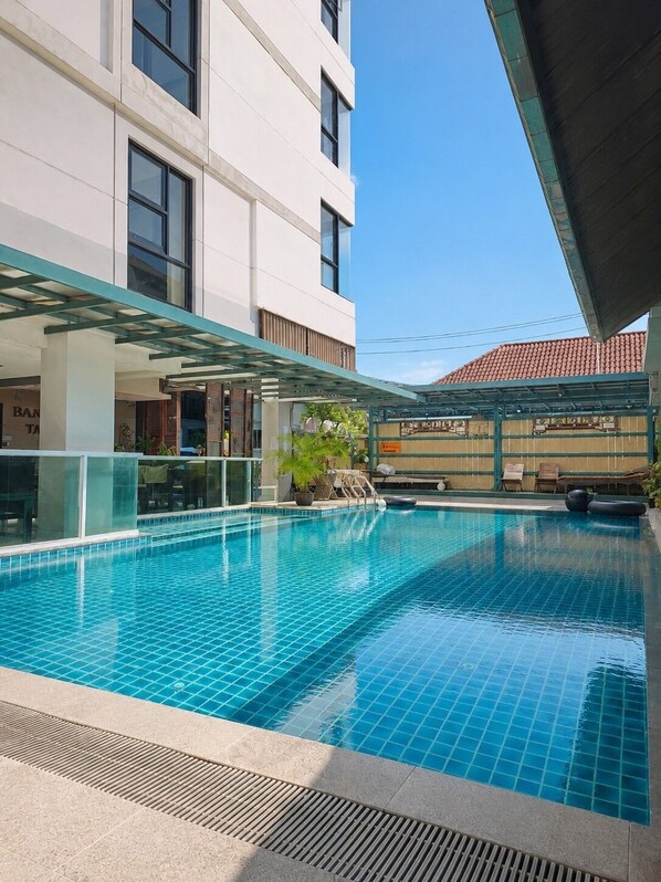A swimming pool with a tiled deck, a covered seating area, and a building with large windows in the background. - Baan Tanwa - MRT Ratchadapisek (Bangkok)
