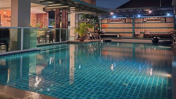 Una piscina con solárium empedrado, una zona techada con asientos y un edificio con amplias ventanas.
