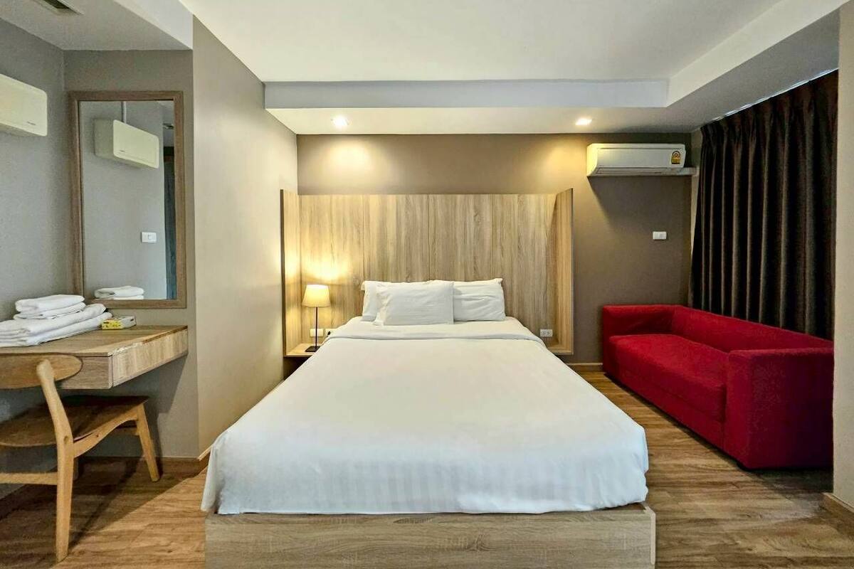 A hotel room with a large bed, a desk with a chair, a red sofa, and an air conditioning unit on the wall.