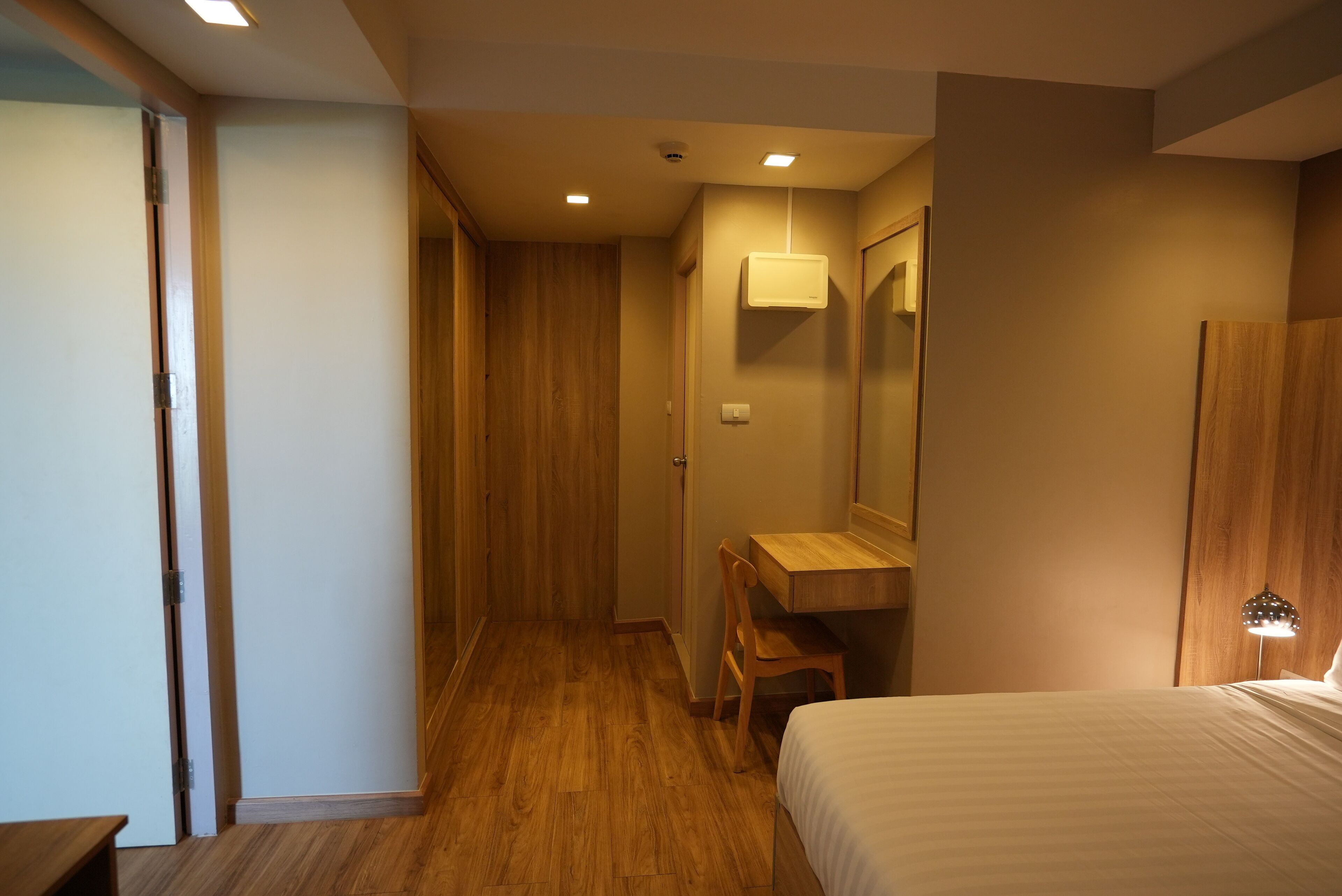 A hotel room with a wooden floor, a bed, a desk with a chair, a mirror, and a wardrobe.