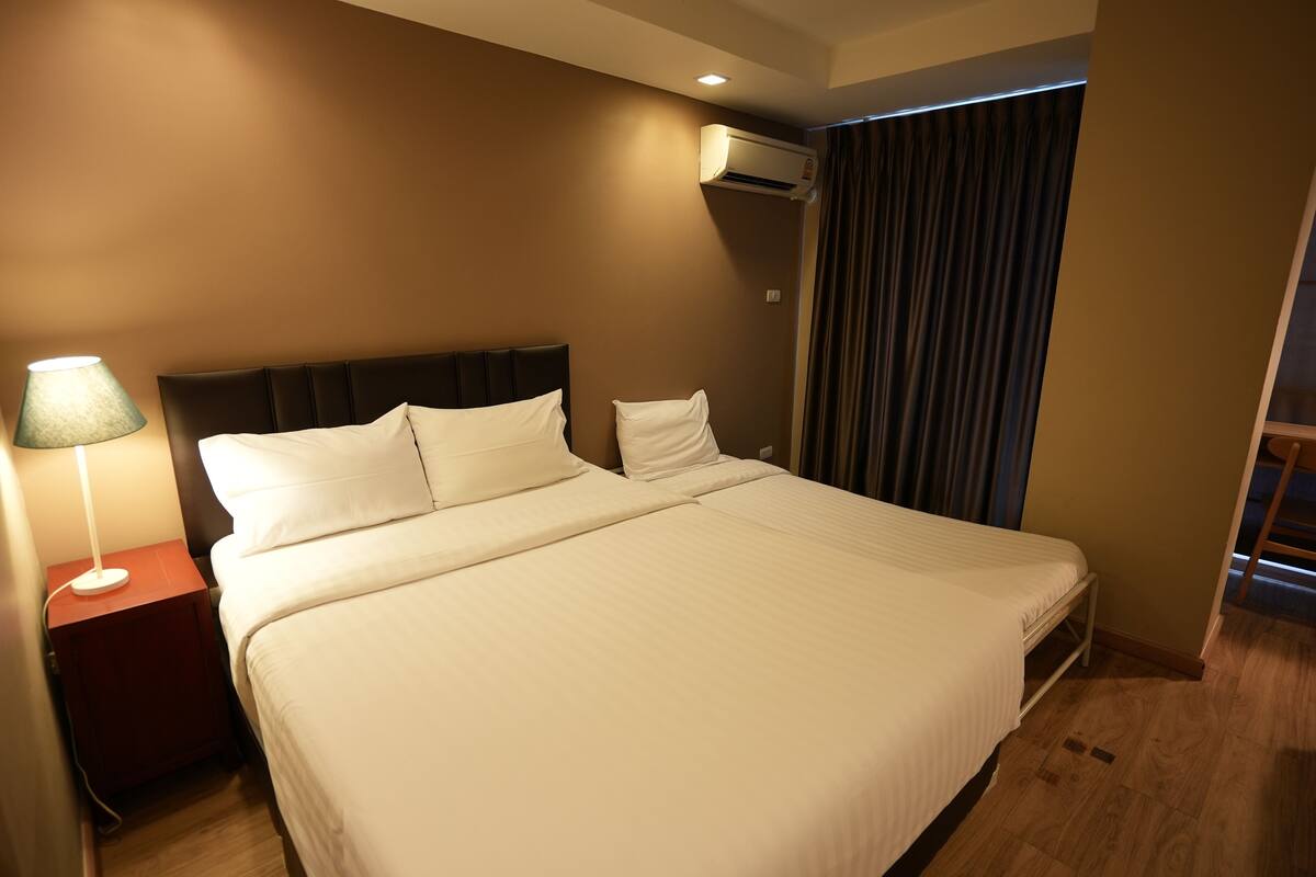 A hotel room with a large bed, a bedside lamp, an air conditioning unit, and a view of the outside through a window with curtains.