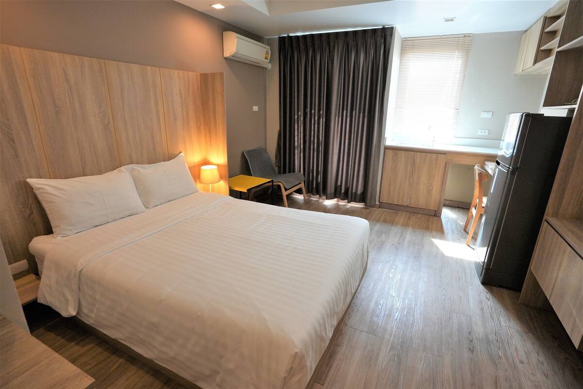 A hotel room with a large bed, a desk, a chair, a refrigerator, and a window with curtains.