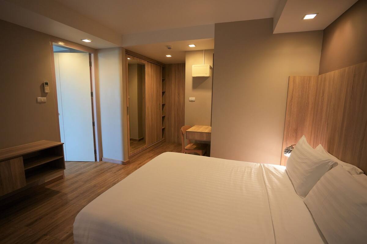 A modern hotel room with a large bed, wooden flooring, a built-in wardrobe, a small desk, and a bedside table.
