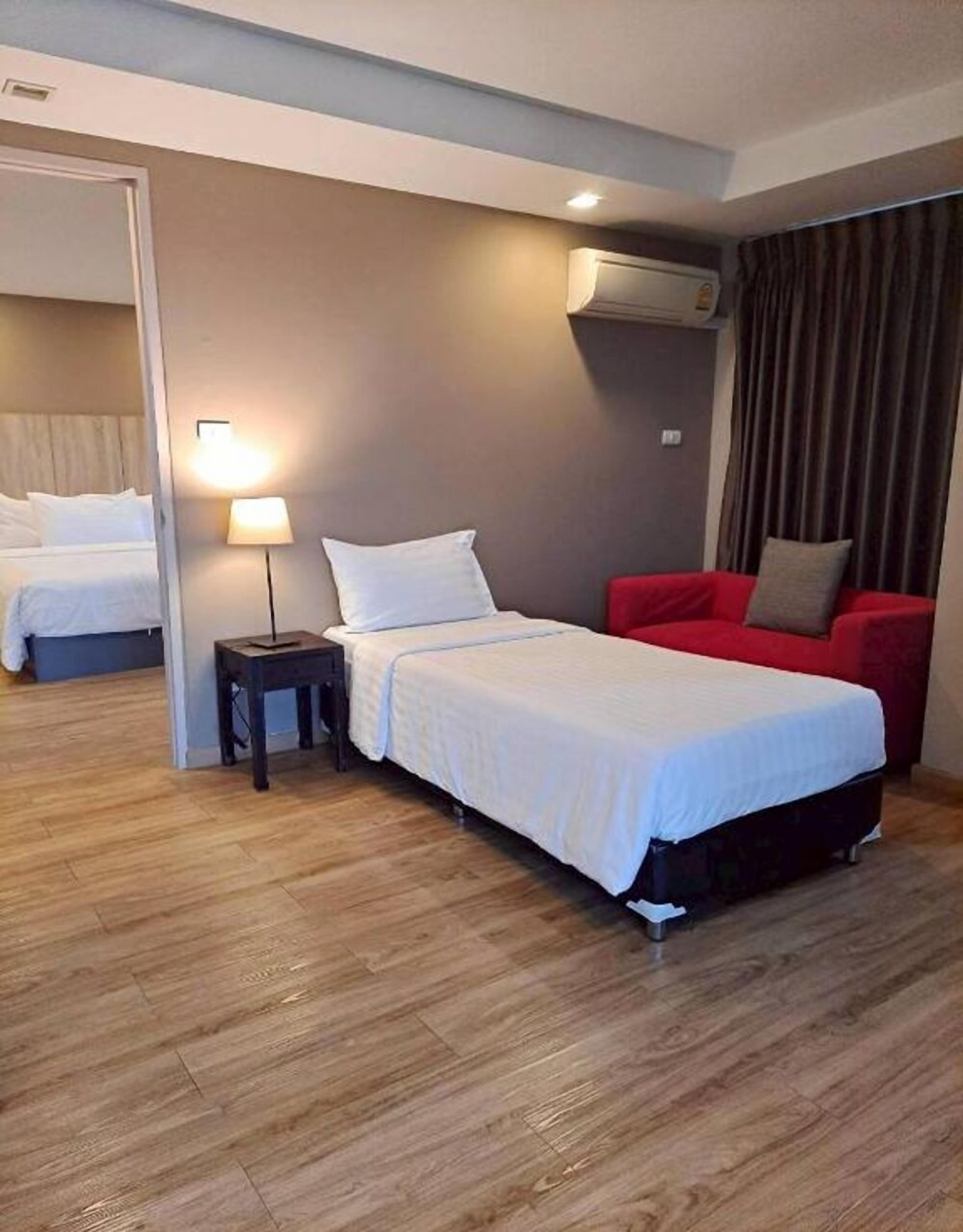 A hotel room with a single bed, a red armchair, a bedside table with a lamp, and an air conditioning unit on the wall.