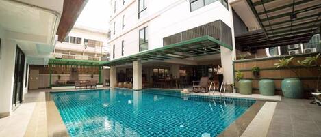 An indoor swimming pool with a tiled floor, surrounded by a covered seating area and a building with multiple floors and windows.