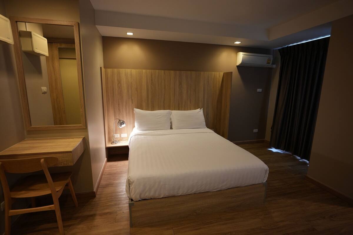 A hotel room with a wooden desk, a bed with white bedding, a nightstand with a lamp, and an air conditioning unit.