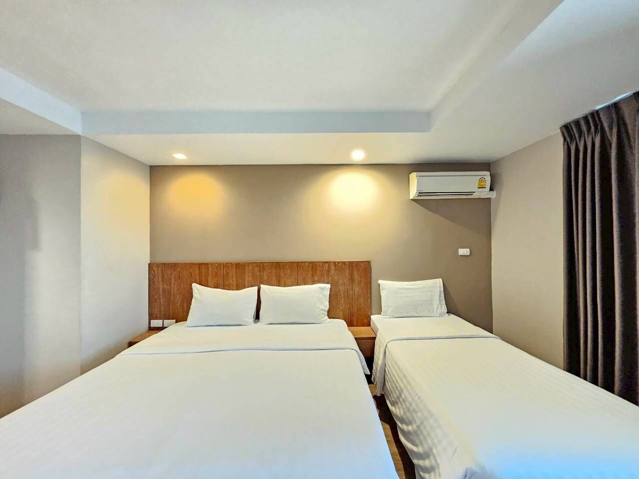 Habitación de hotel con dos camas, cabecera de madera, unidad de aire acondicionado y una cortina a la derecha.