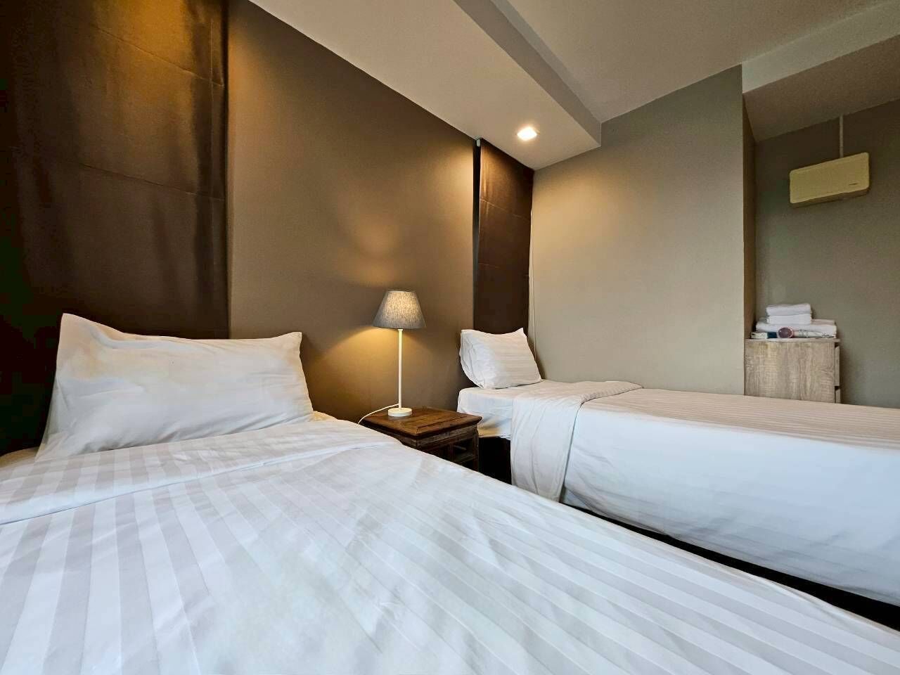 A hotel room with two beds, a bedside table with a lamp, and a small wooden side table with towels.
