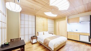 Una habitación de hotel moderna con suelo de madera, una cama con ropa blanca, una mesita de noche pequeña, un escritorio con una silla y una kitchenette con un lavamanos y un refrigerador.