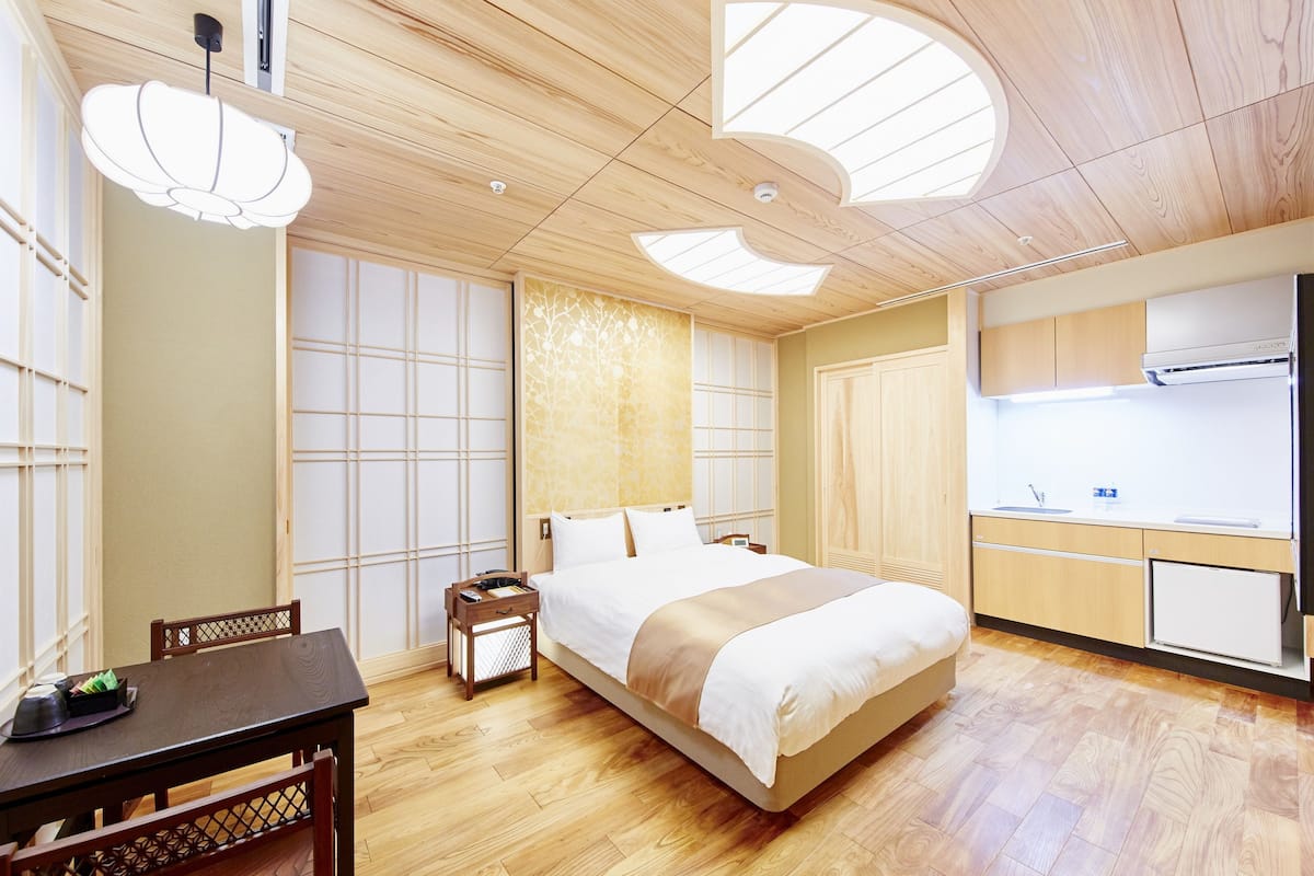 A modern hotel room with a wooden floor, a bed with white bedding, a small bedside table, a desk with a chair, and a kitchenette with a sink and a refrigerator.