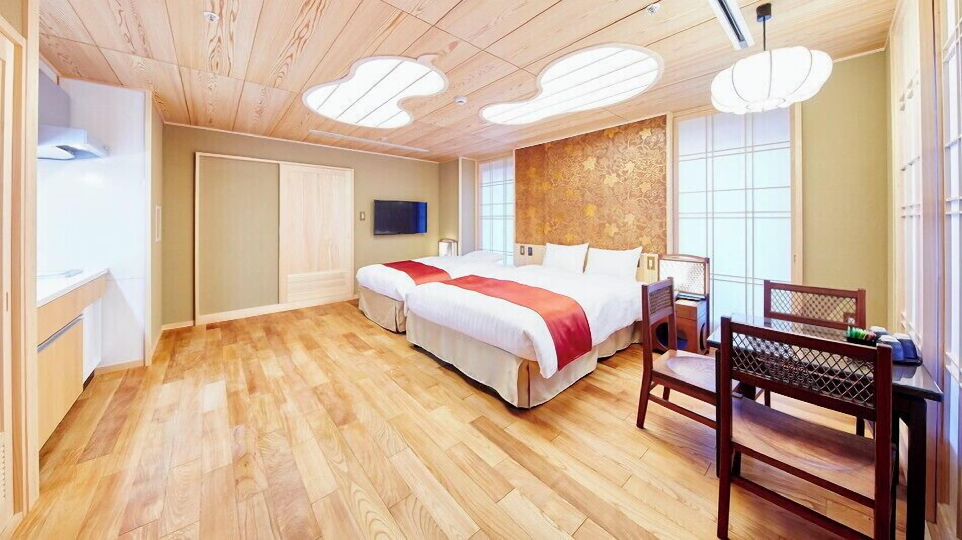 A bedroom with a wooden floor, a bed with a red and white bedspread, a wooden chair, and a TV mounted on the wall.