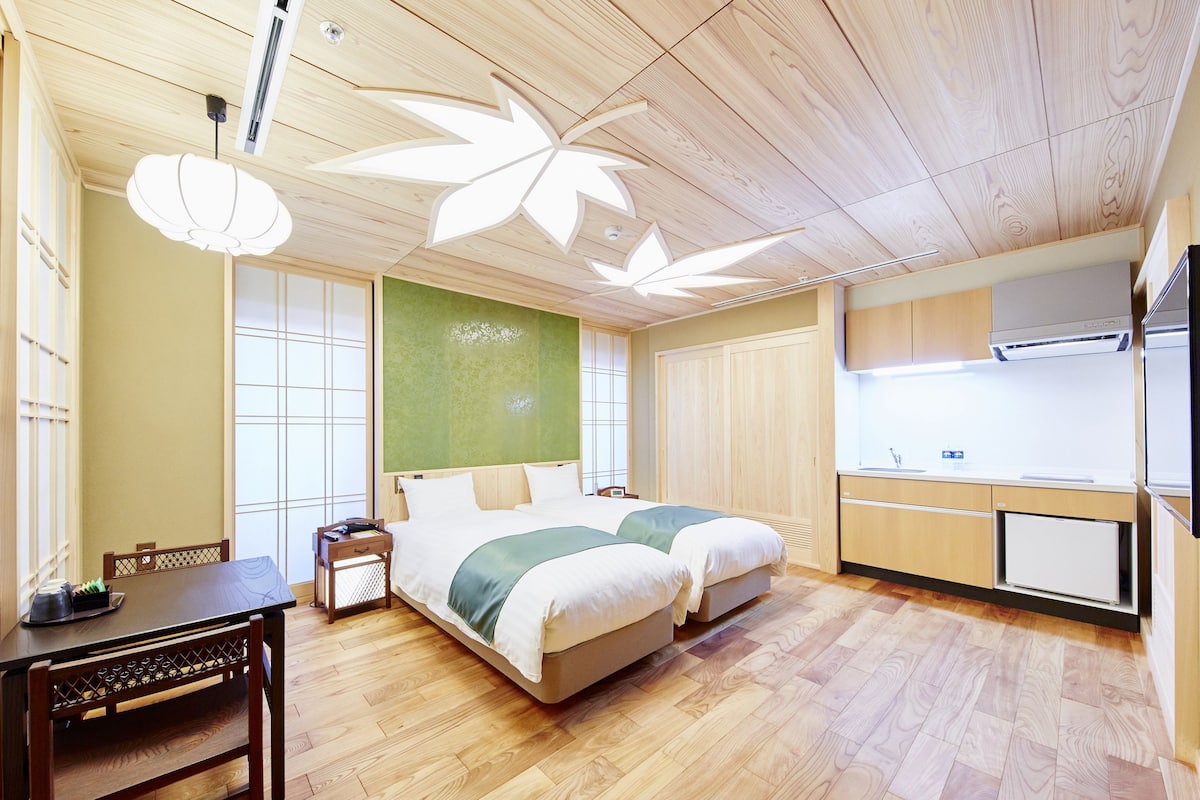 A hotel room with a wooden ceiling, a bed with a green blanket, a small table with a chair, and a kitchenette area.
