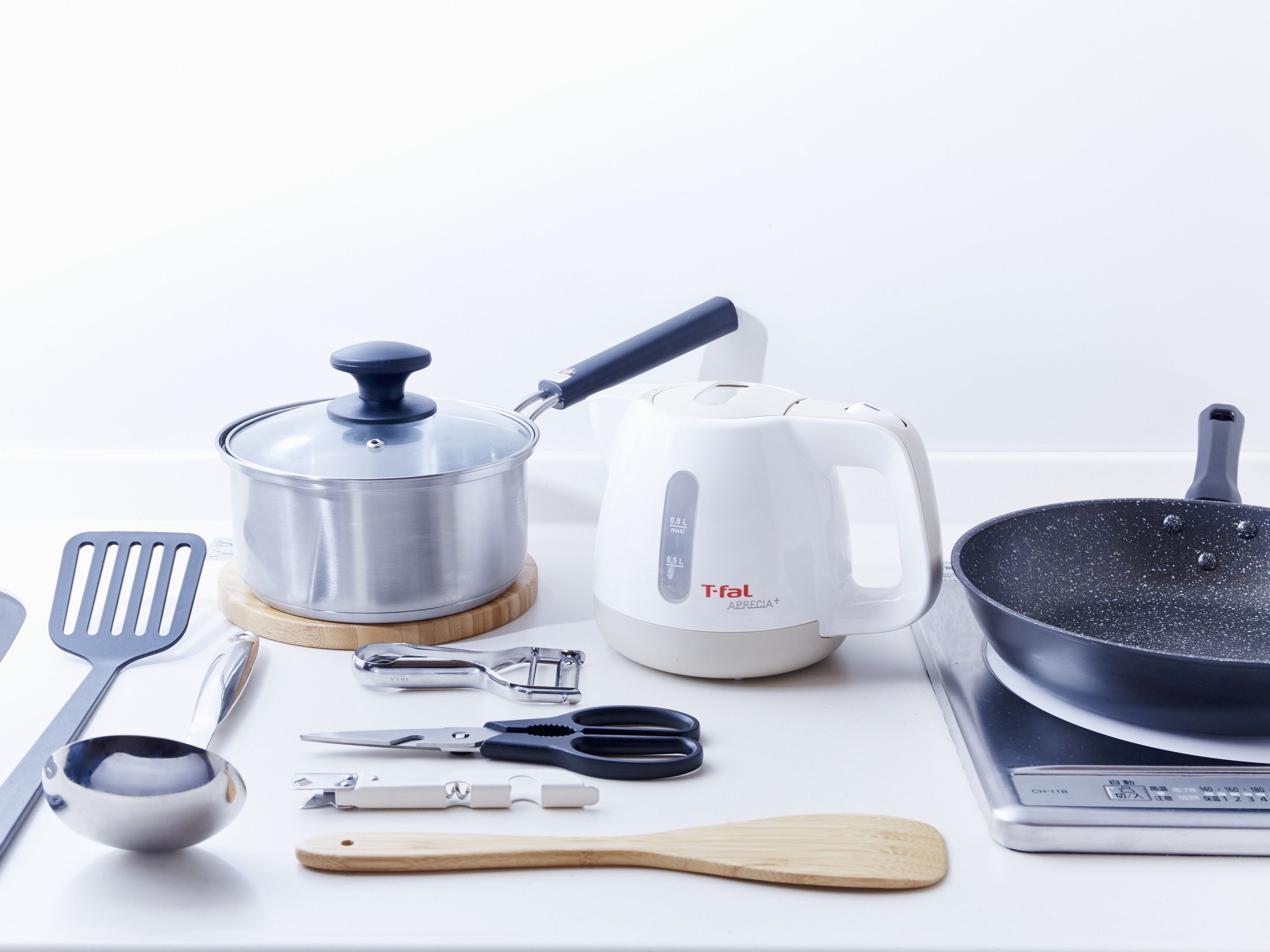 A kitchen counter with a stainless steel pot, a white electric kettle, a frying pan, a wooden spatula, a metal spatula, a metal measuring cup, a pair of scissors, and a metal can opener.