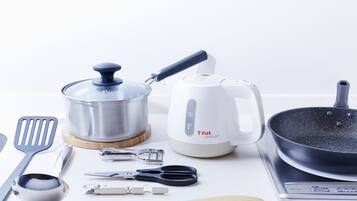 A kitchen counter with a stainless steel pot, a white electric kettle, a frying pan, a wooden spatula, a metal spatula, a metal measuring cup, a pair of scissors, and a metal can opener.