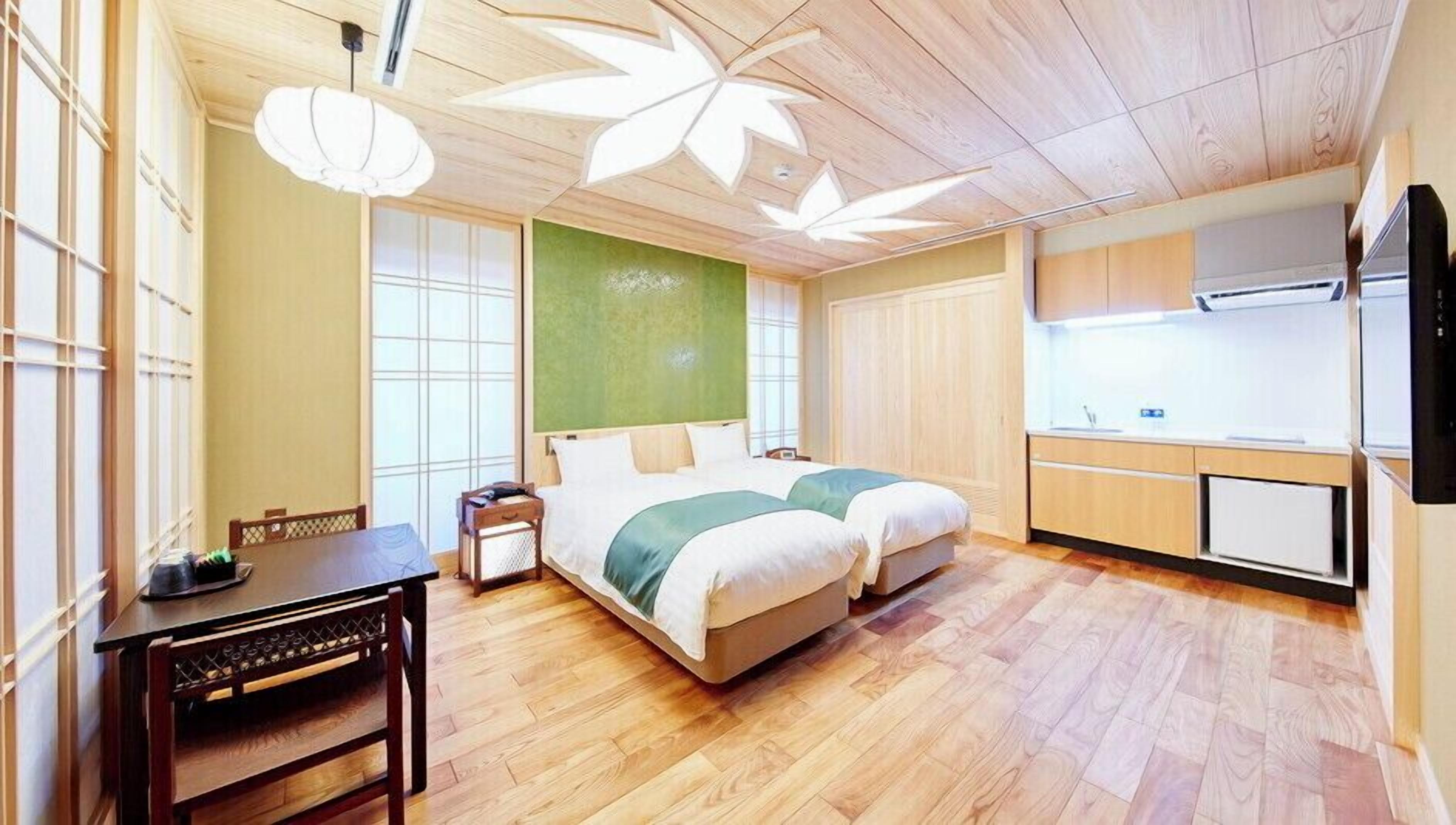 A hotel room with a bed, a desk, a chair, a small table, a window, and a kitchenette.
