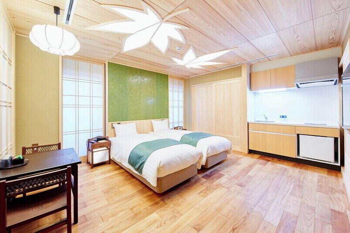 A hotel room with a bed, a desk, a chair, a small table, a window, and a kitchenette.