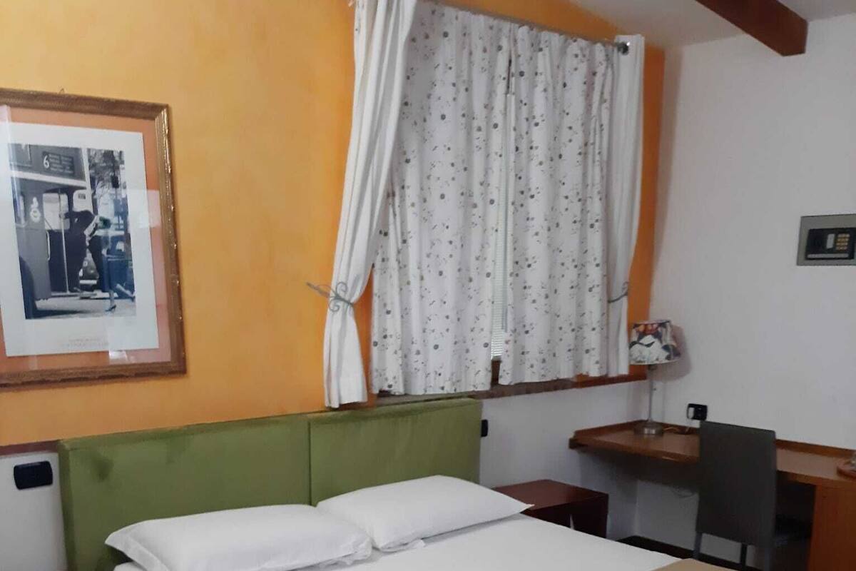 A single-bed room with a green headboard, a desk with a chair, a window with floral curtains, and a framed picture on the wall.