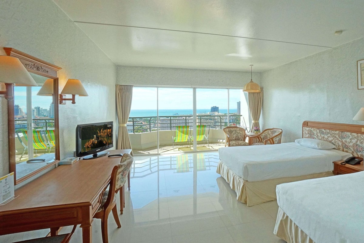 Une chambre d’hôtel avec un balcon, un bureau avec une télévision, deux lits et une chaise.