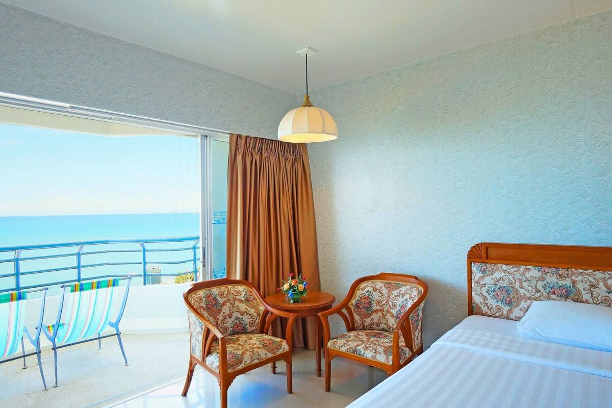 Une chambre d’hôtel avec un lit, deux chaises, une petite table avec un vase, un balcon avec une balustrade et une vue sur la mer.
