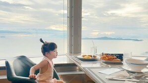 Uma criança sentada à mesa comendo, com vista para a água e terra ao longe.