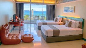 Quarto de hotel com duas camas, uma escrivaninha, uma cadeira e um sofá. Há uma varanda com vista para o mar e para as montanhas.