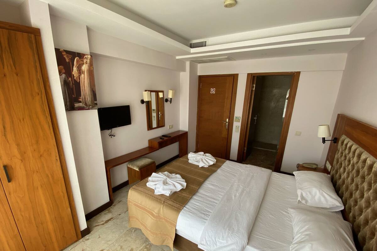 A hotel room with a bed, bedside tables, a desk, a mirror, a television, and a door.