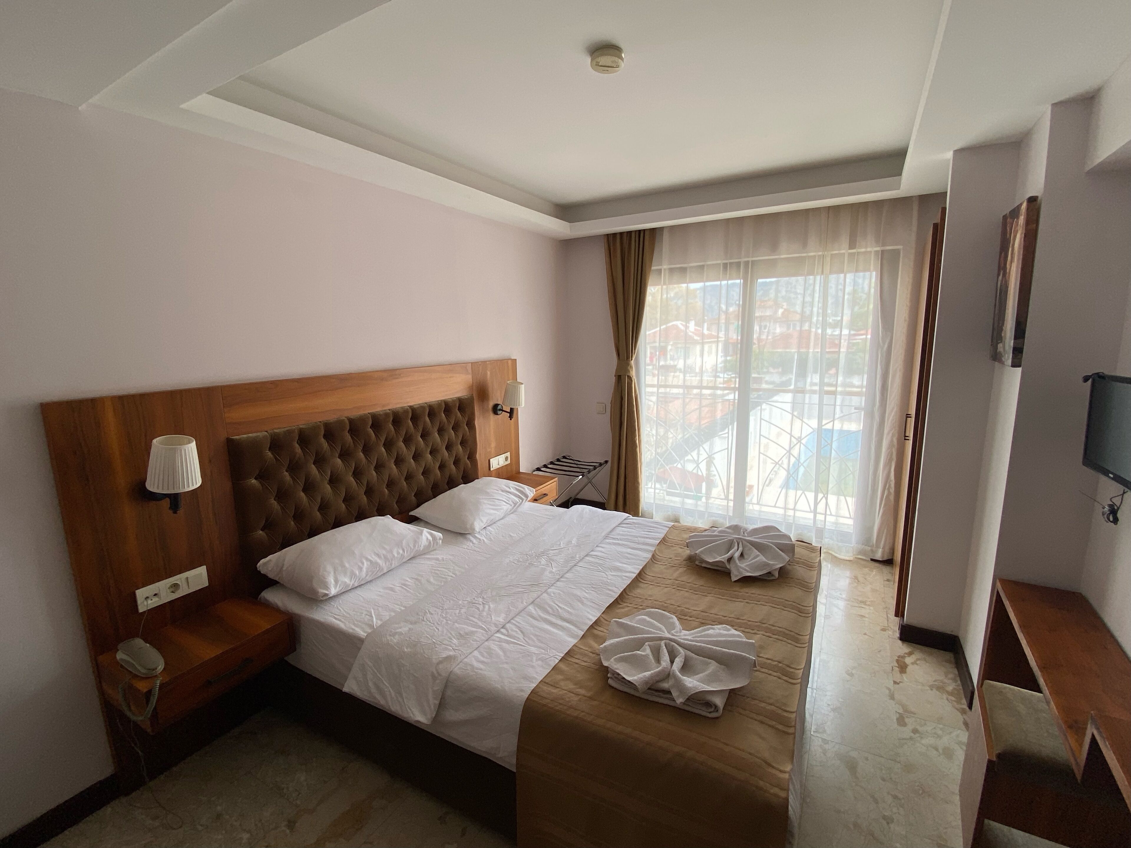A hotel room with a large bed, a wooden headboard, a bedside table with a lamp, a TV mounted on the wall, and a window with curtains.
