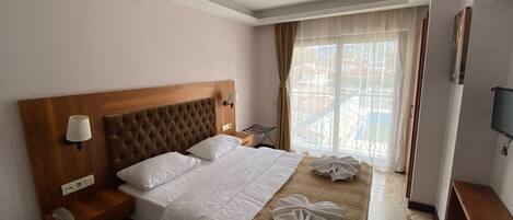 A hotel room with a large bed, a wooden headboard, a bedside table with a lamp, a TV mounted on the wall, and a window with curtains.