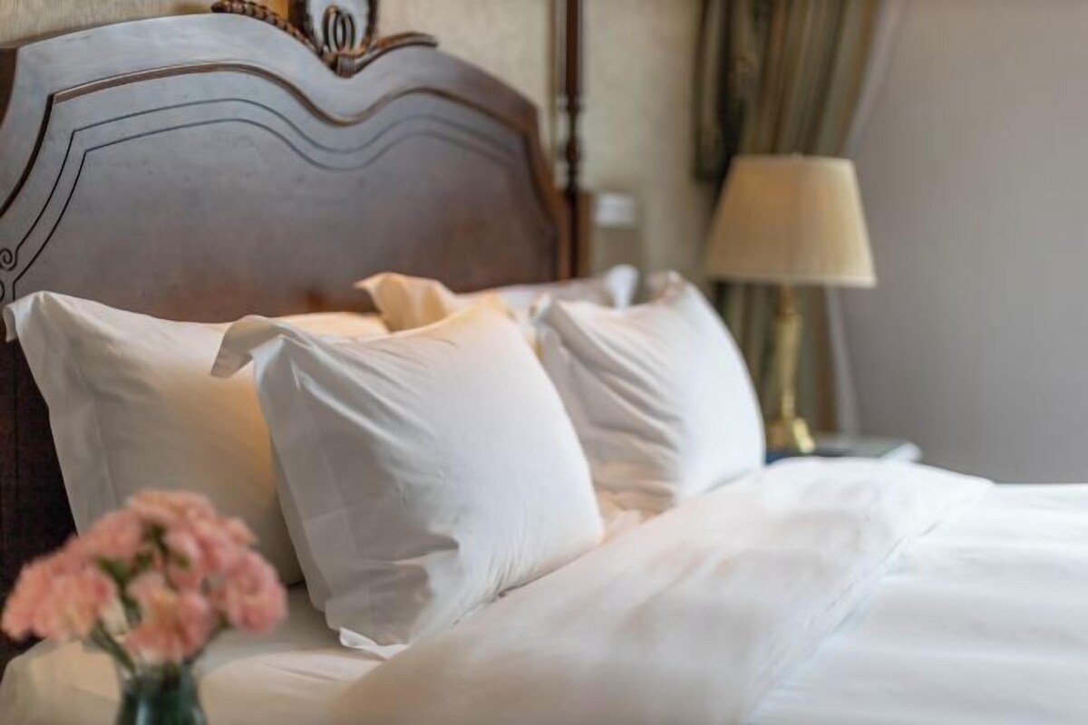 A neatly made bed with white linens and pillows, a wooden headboard, and a bedside lamp.