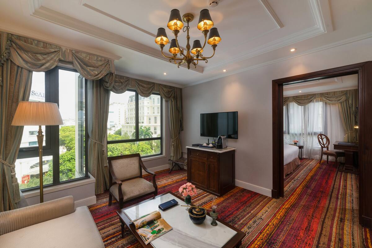 A hotel room with a large window, a television, a desk, a chair, and a patterned carpet.