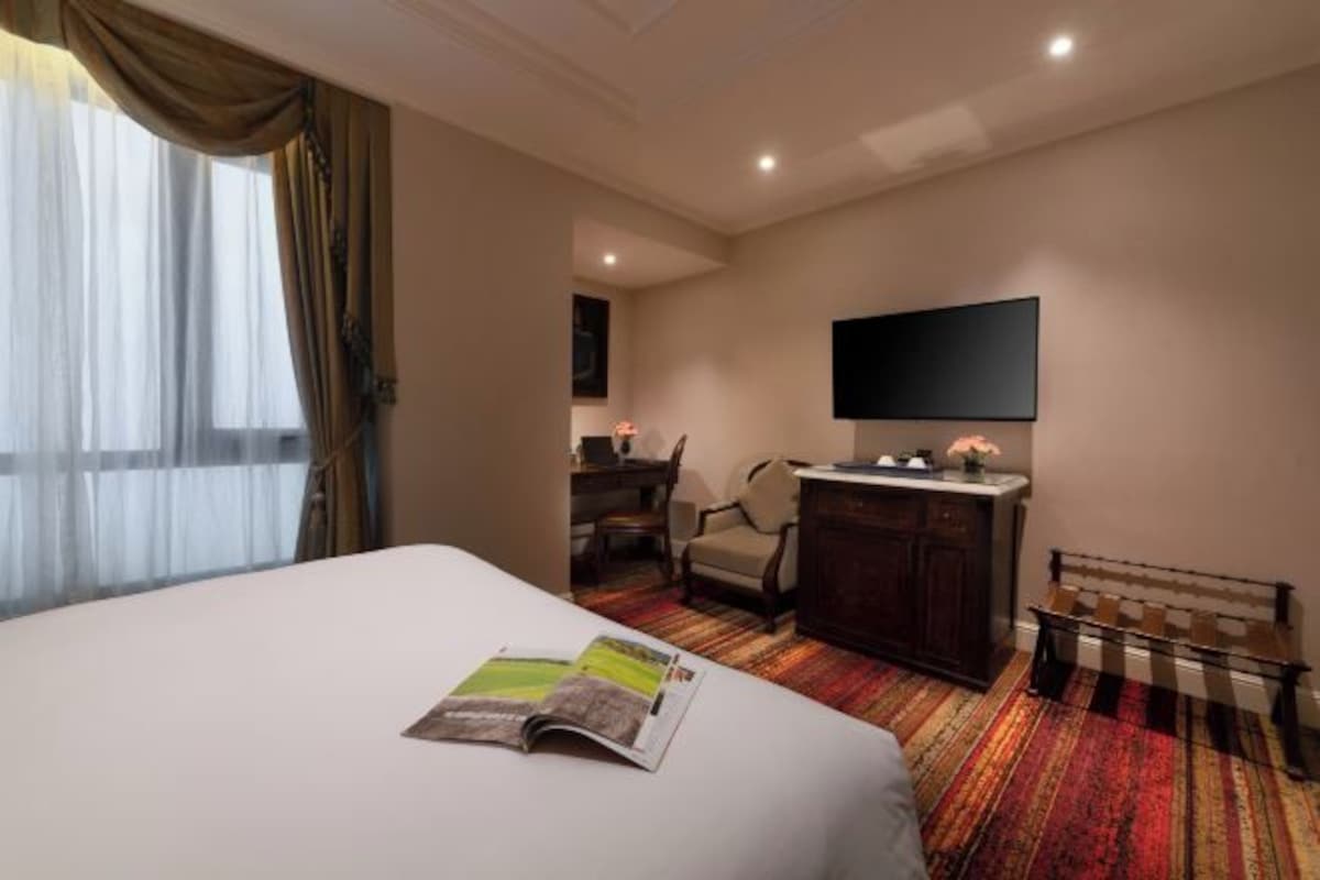 A hotel room with a large bed, a desk with a chair, a television, and a bench.