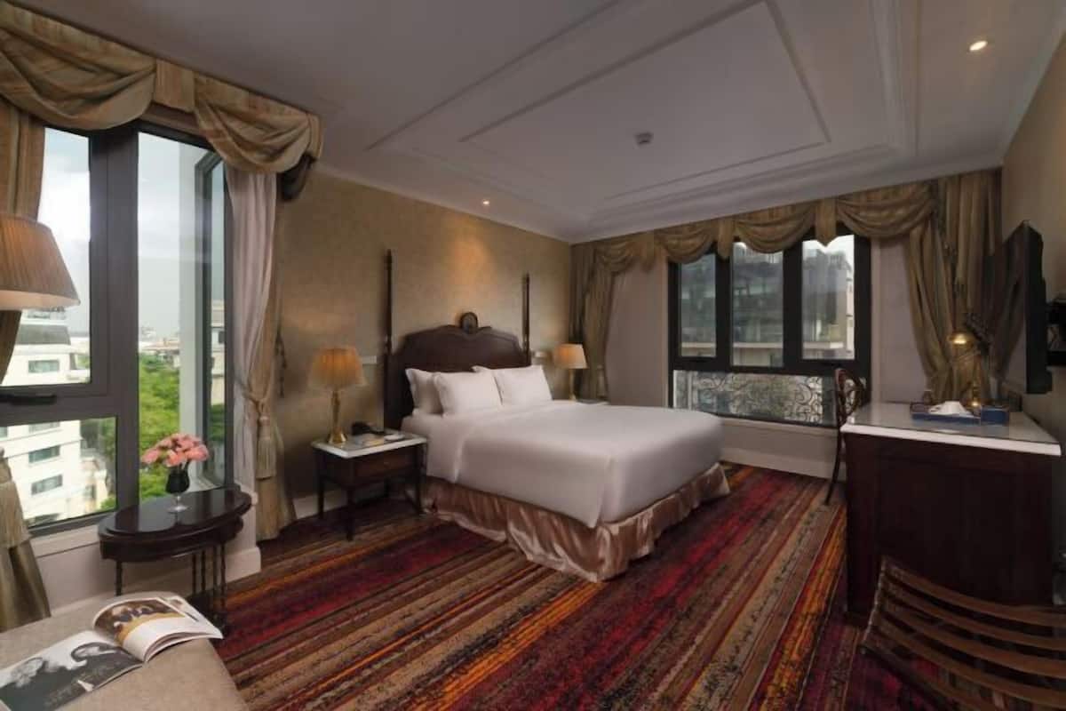 A hotel room with a large bed, a desk, a chair, a window with curtains, and a view of buildings outside.