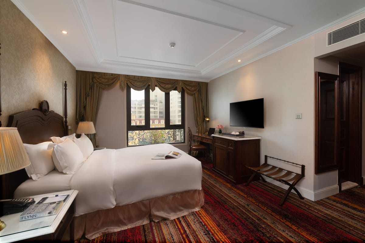 A hotel room with a large bed, a desk, a television, and a window with a view of buildings.