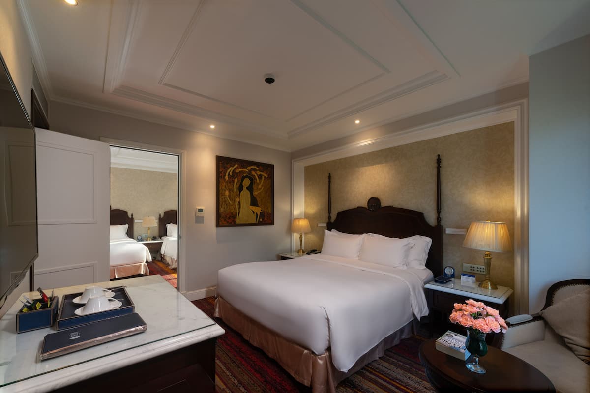 A hotel room with a large bed, bedside tables, a desk with a chair, a mirror, and a painting on the wall.