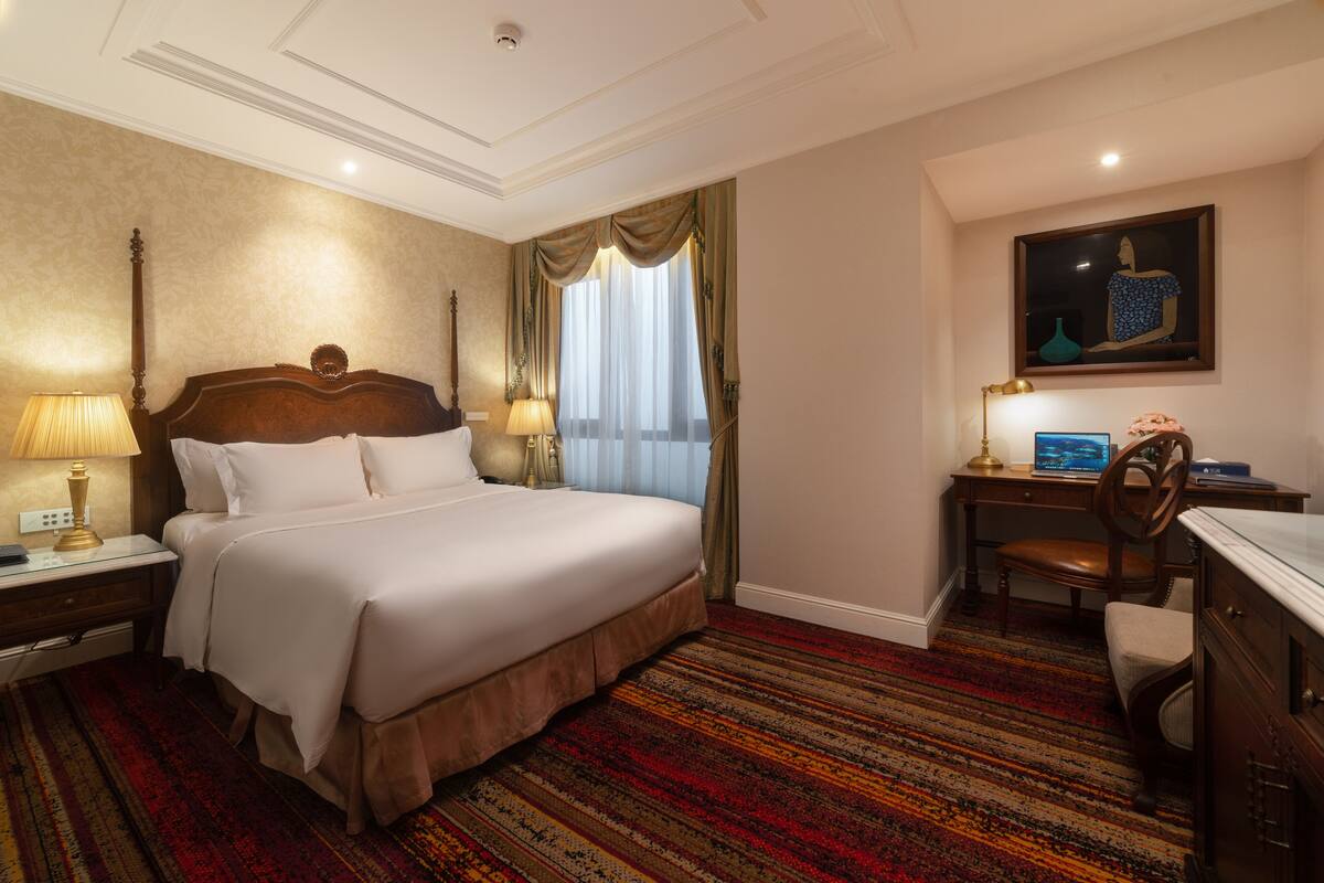 A hotel room with a large bed, a desk with a computer, a chair, and a desk lamp.