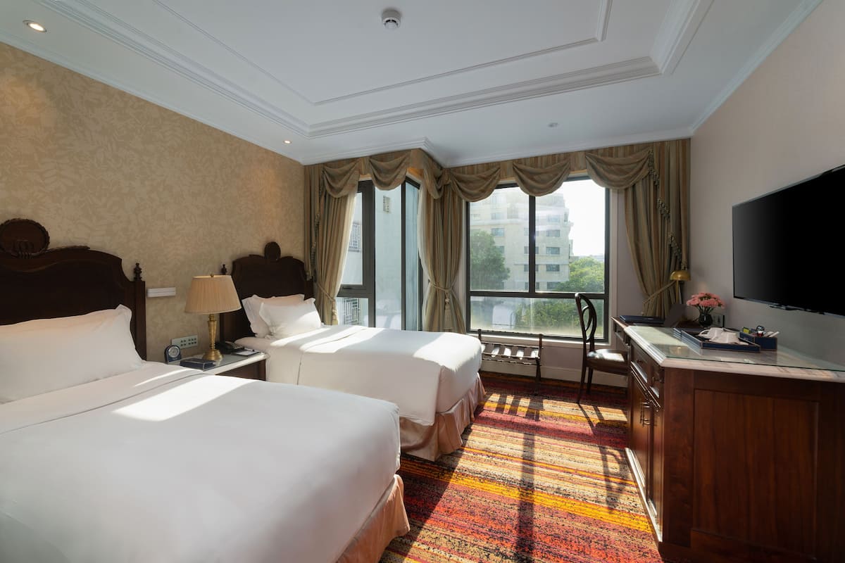 A hotel room with two beds, a desk with a TV, and large windows with curtains.