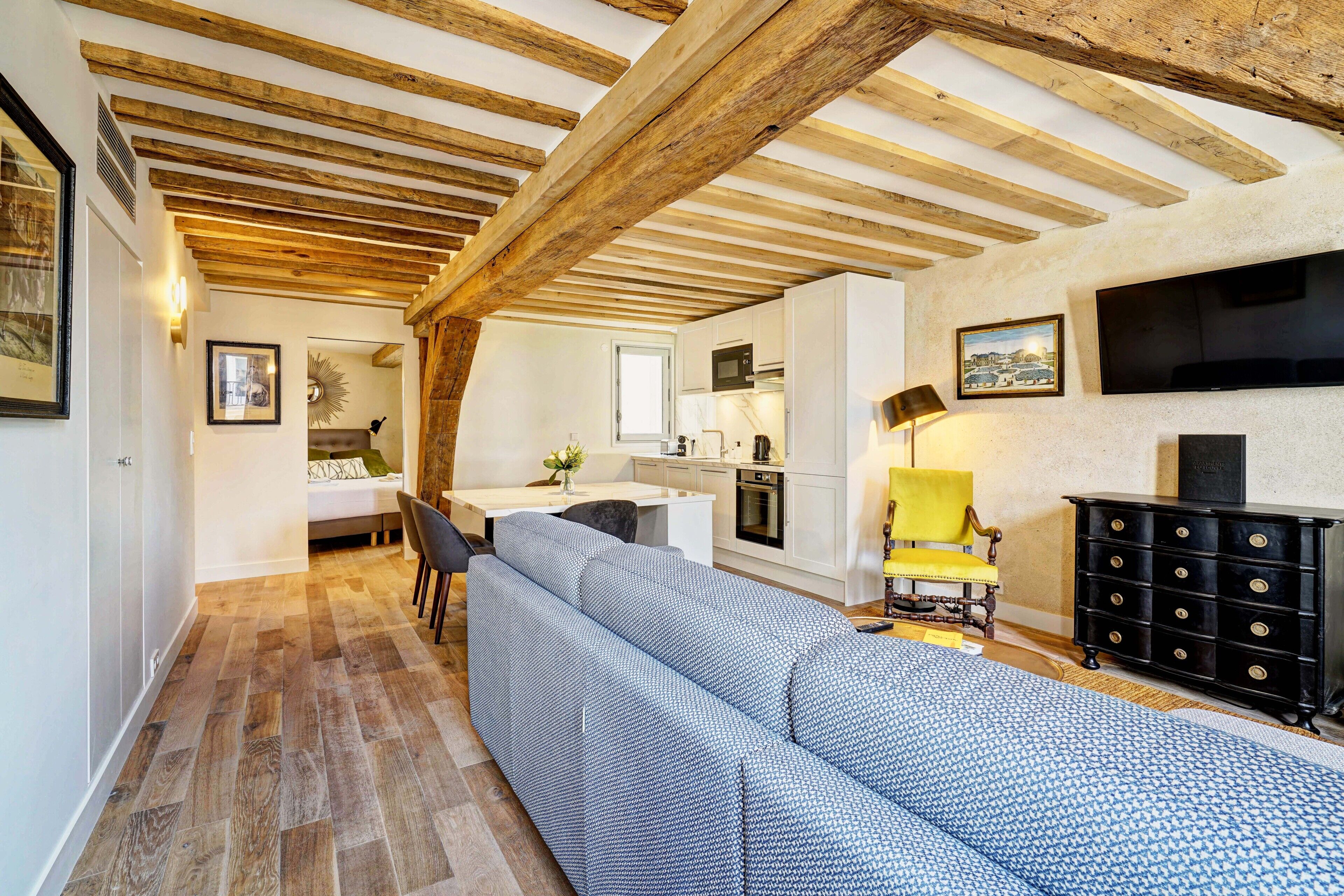 A modern living room with a large sofa, wooden beams on the ceiling, a kitchen area, and a TV mounted on the wall.