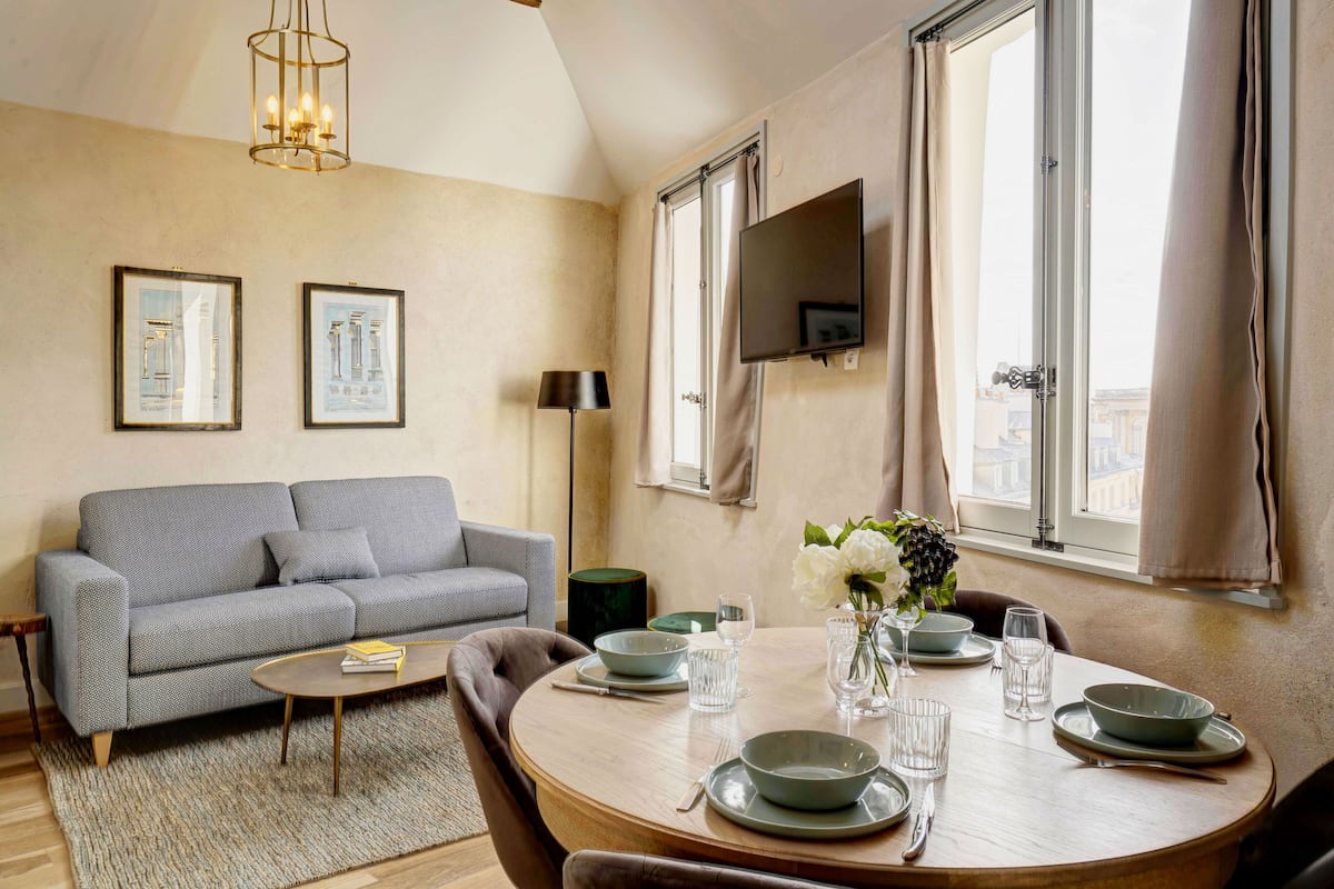A living room with a sofa, coffee table, dining table set with plates and glasses, a flat-screen TV mounted on the wall, and a window with curtains.