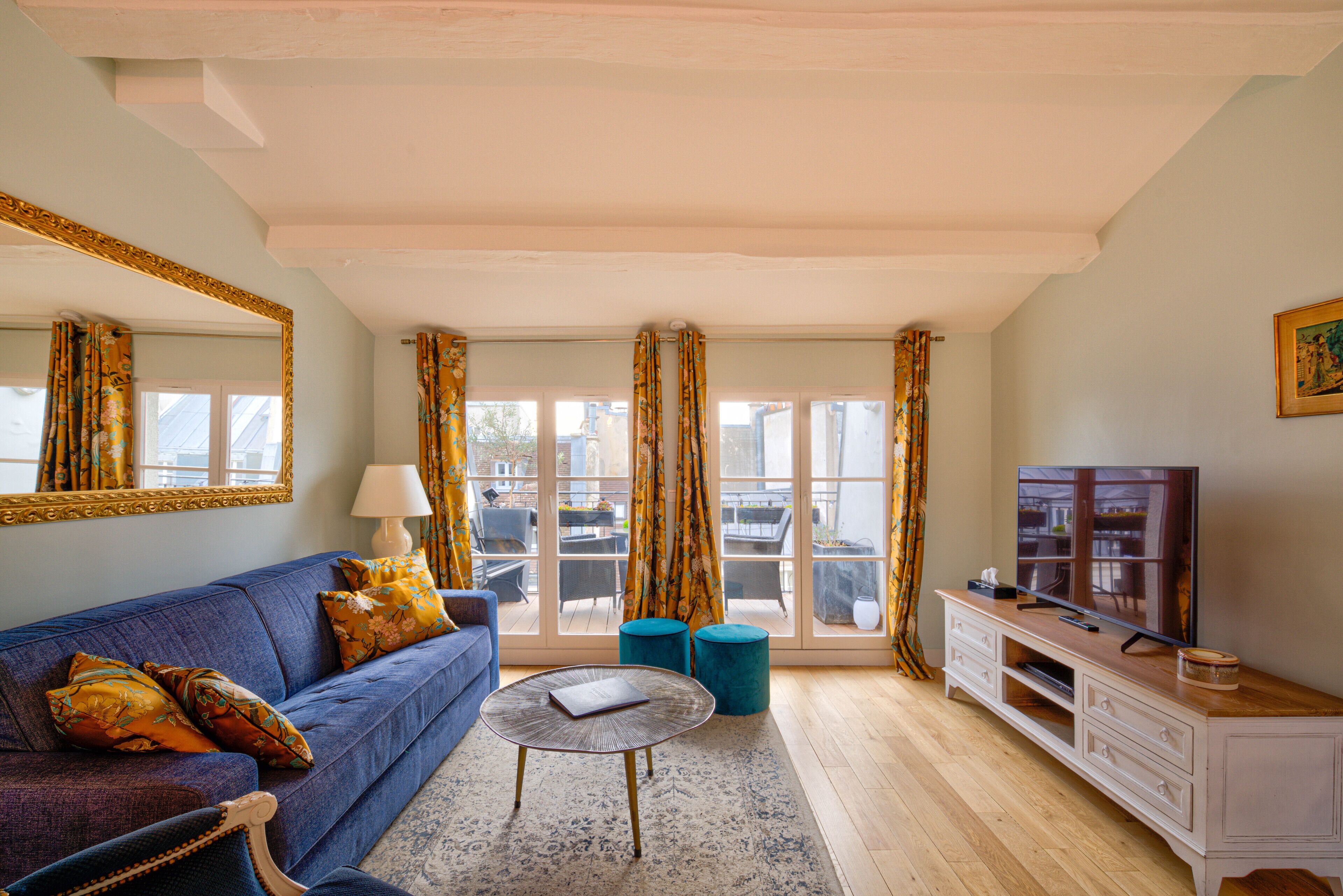 A living room with a blue sofa, a round coffee table, a TV stand, and a large window with curtains.