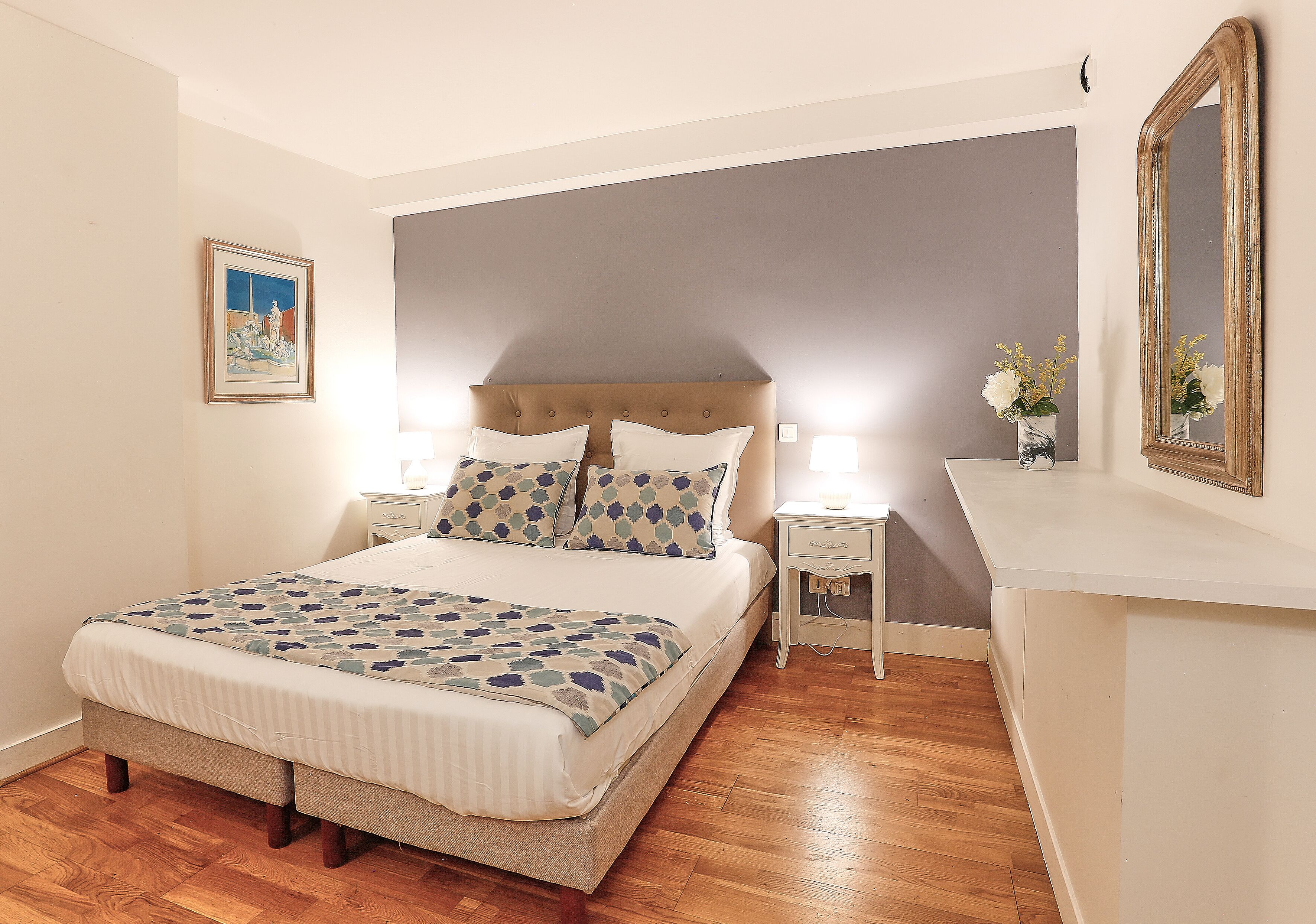 Un dormitorio con cama, mesitas de noche, tocador, espejo y un cuadro en la pared.