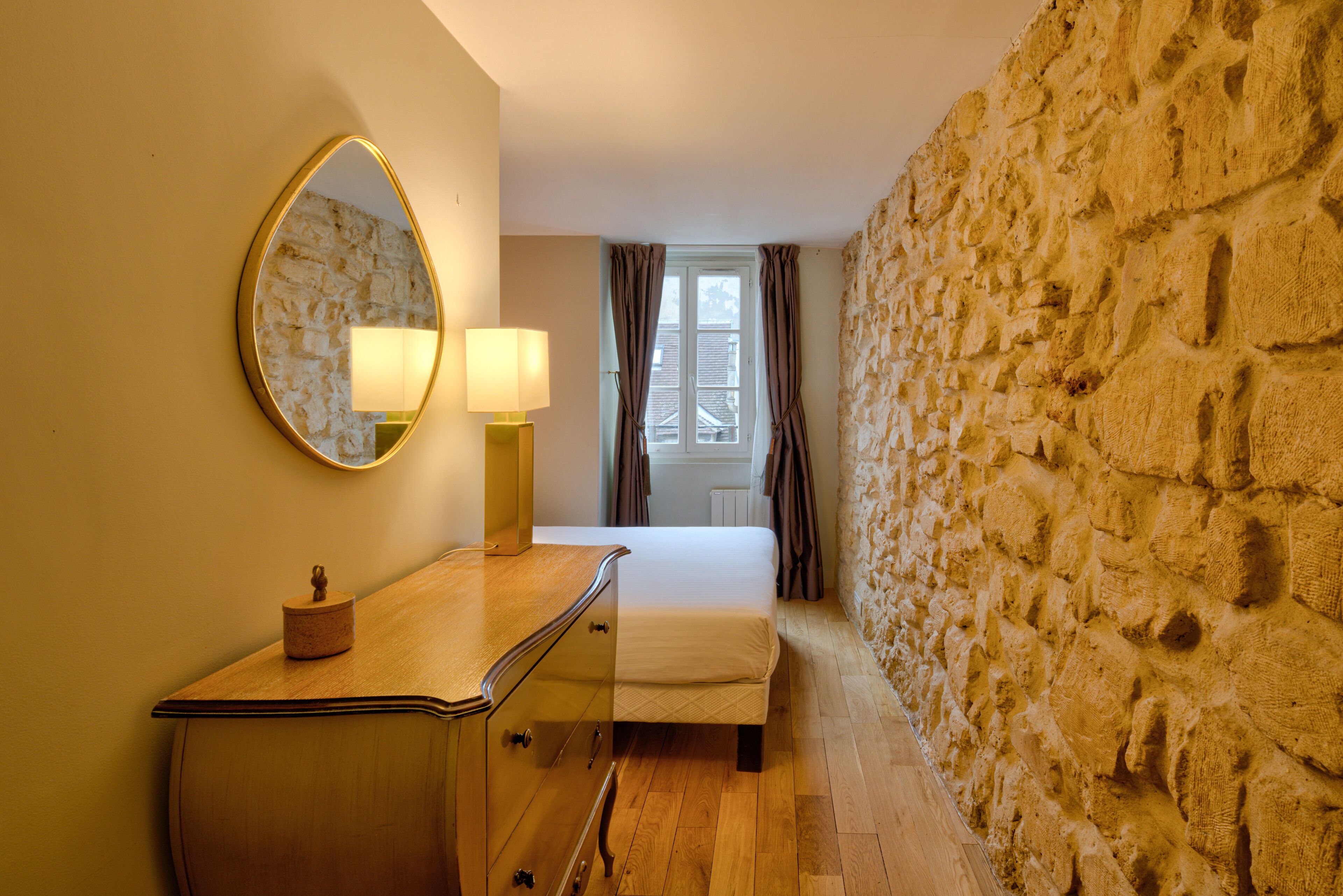 A bedroom with a stone wall, a bed, a dresser, and a mirror.