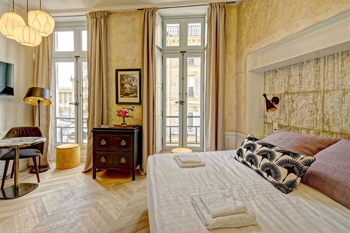 A hotel room with a large bed, a desk with a chair, a dresser, and a large window with curtains.