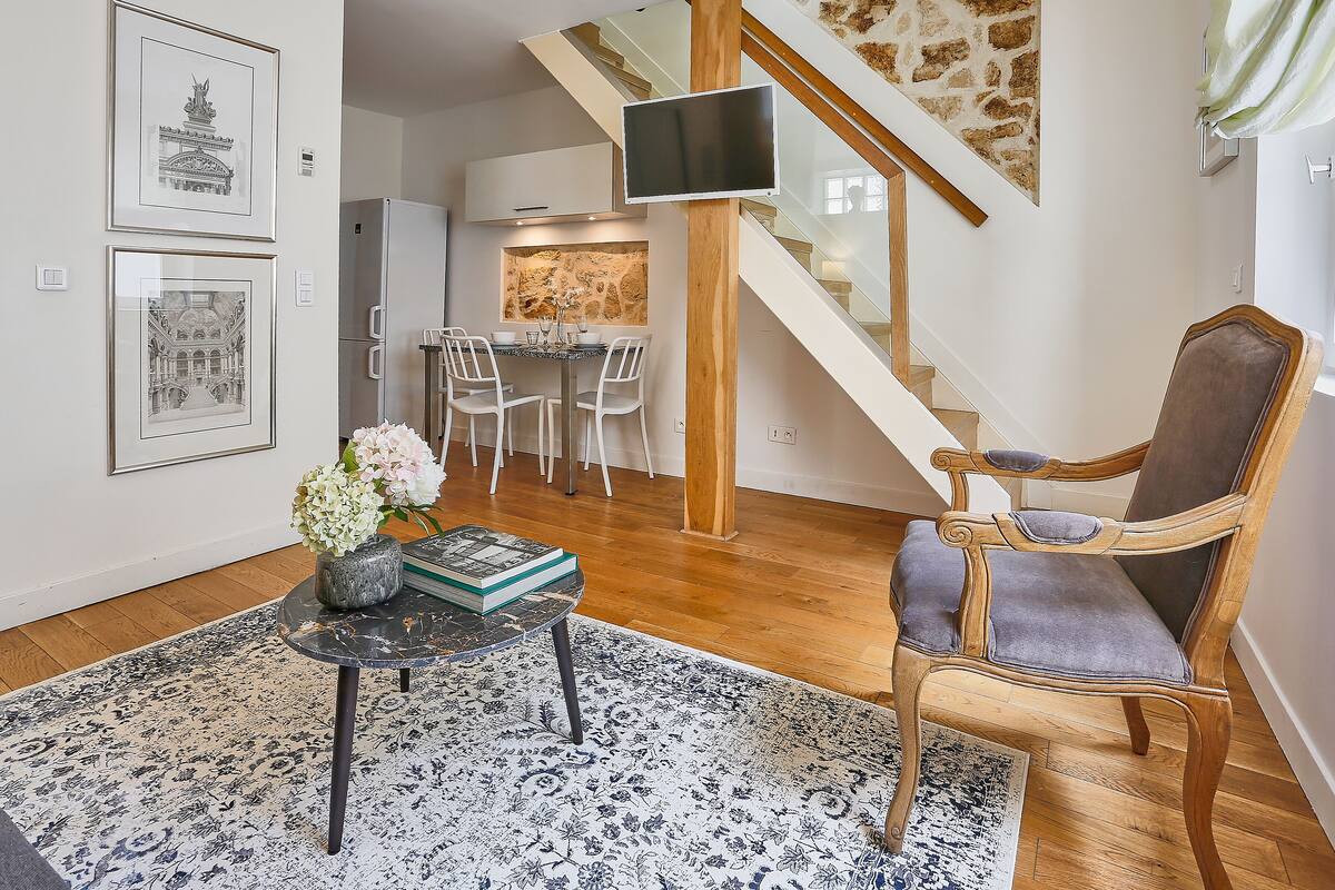 A modern living room with a wooden chair, a round table with a vase of flowers, and a staircase with a TV mounted on the wall.