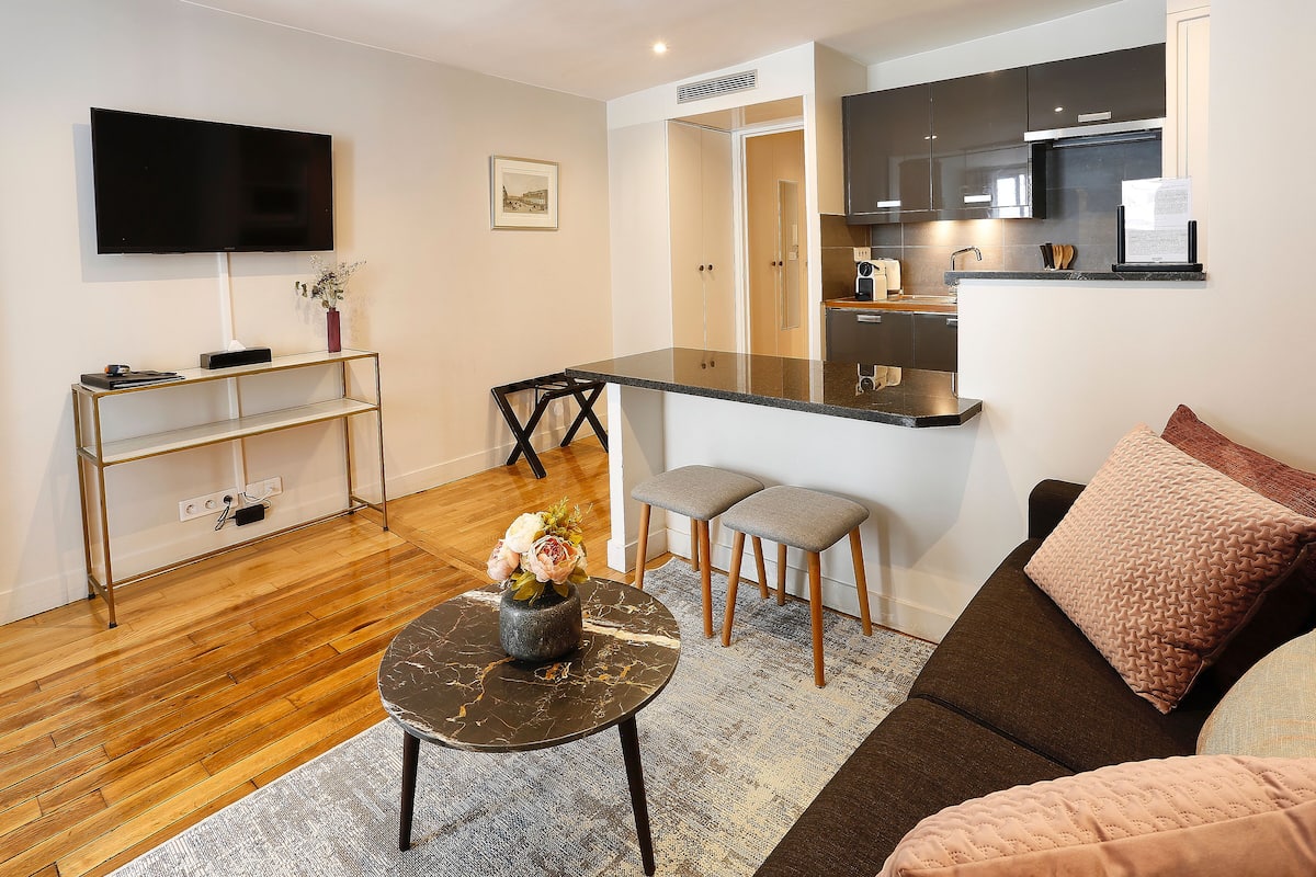 A modern living room with a flat-screen TV, a small round table with a vase, two grey stools, a brown sofa, and a kitchen area with dark cabinets and a microwave.