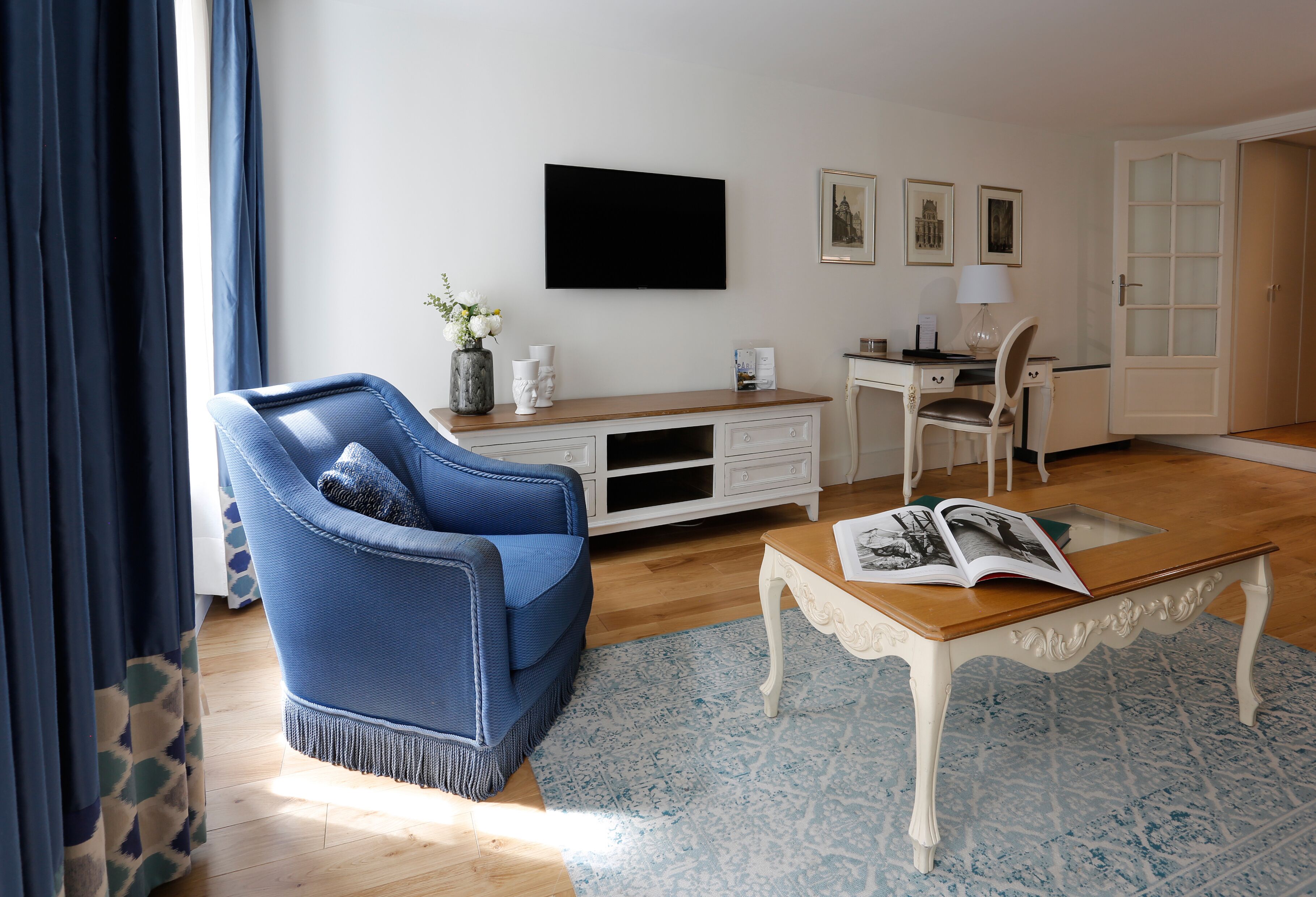 Een woonkamer met een blauwe fauteuil, een houten salontafel met een tijdschrift, een witte tv-meubel en een deur met geëtste glaspanelen.