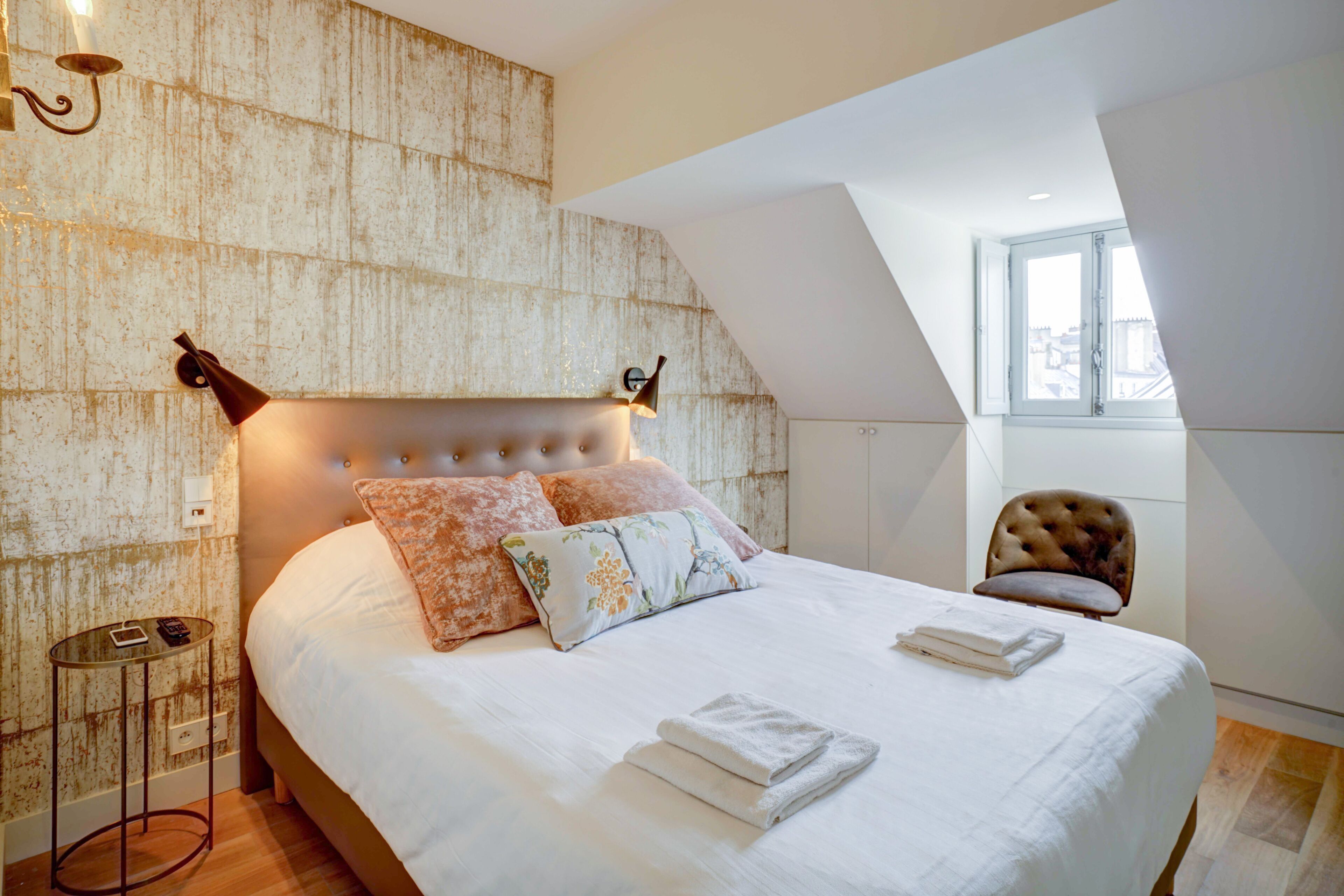 Ein Schlafzimmer mit einem großen Bett, einem Kopfteil, einem Nachttisch, einem Sessel und einem Fenster.