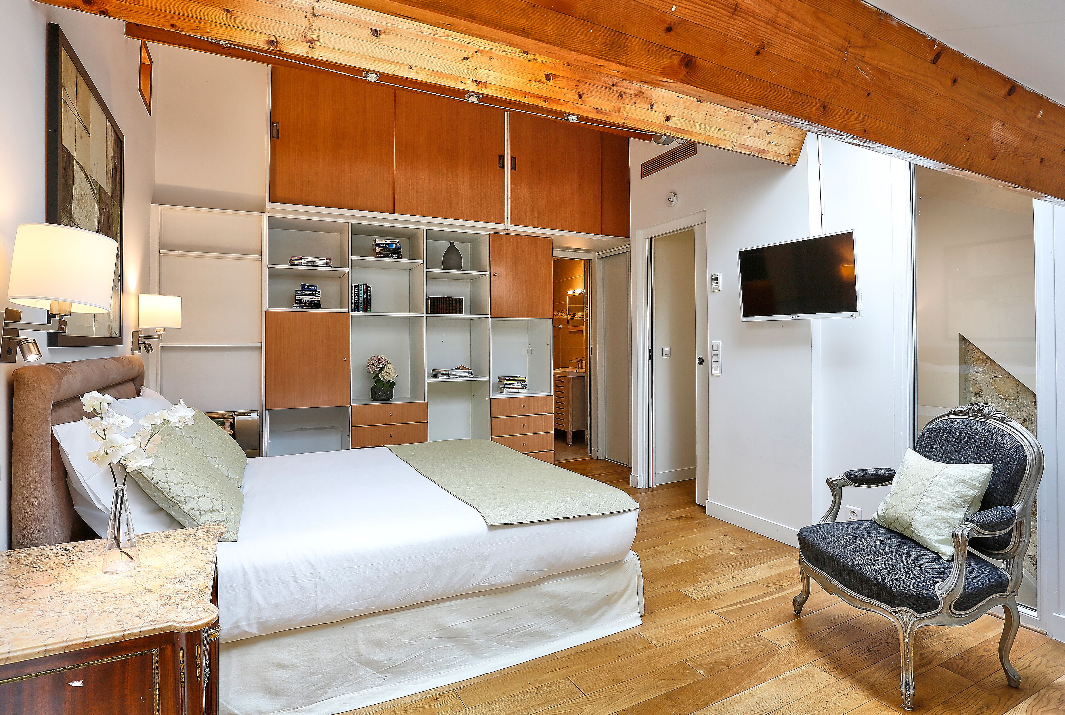 A bedroom with a bed, a chair, a TV, and built-in shelving.
